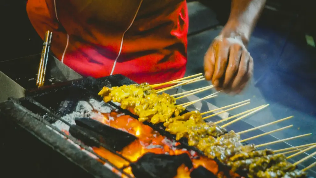 Close-up shot of marinated satay skewers grilling over glowing charcoal embers, highlighting open flames, textured meat, and hands tending the skewers at a night market.