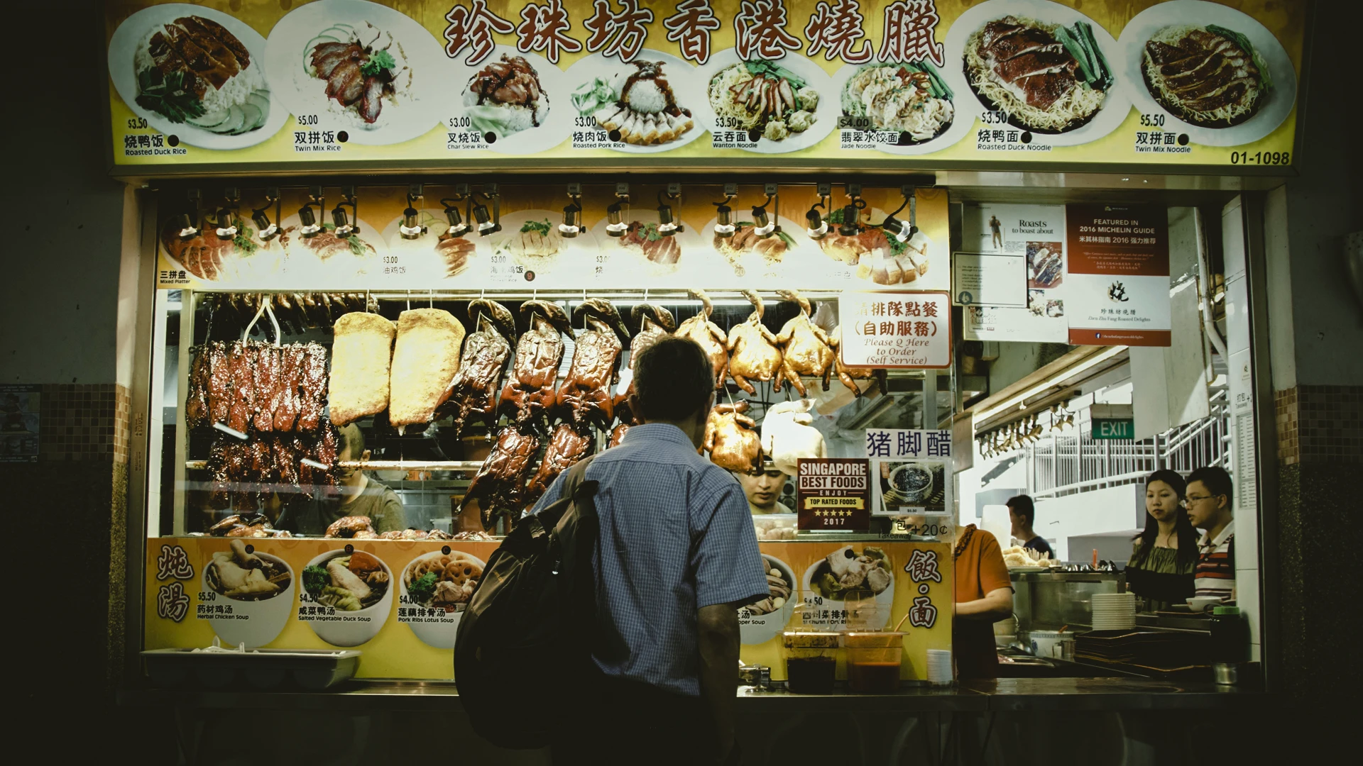 Medium shot of a traditional Chinese roast meat hawker stall, featuring hanging roasted duck and pork beneath a brightly lit menu board with illustrated dishes.