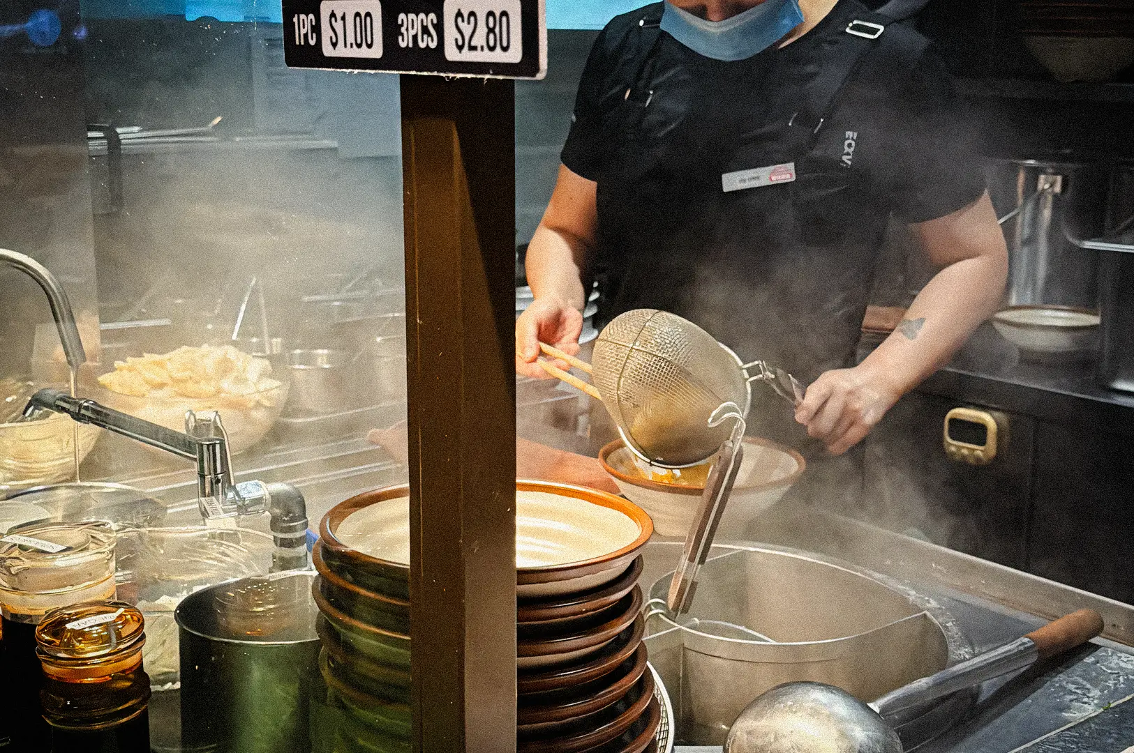 Medium close-up, eye-level shot of a hawker-style kitchen scene showing a cook preparing bak chor mee, steaming noodles being strained over a boiling pot, with stacked ceramic bowls, ladles, and visible steam highlighting live noodle cooking at a Singapore-style noodle stall.
