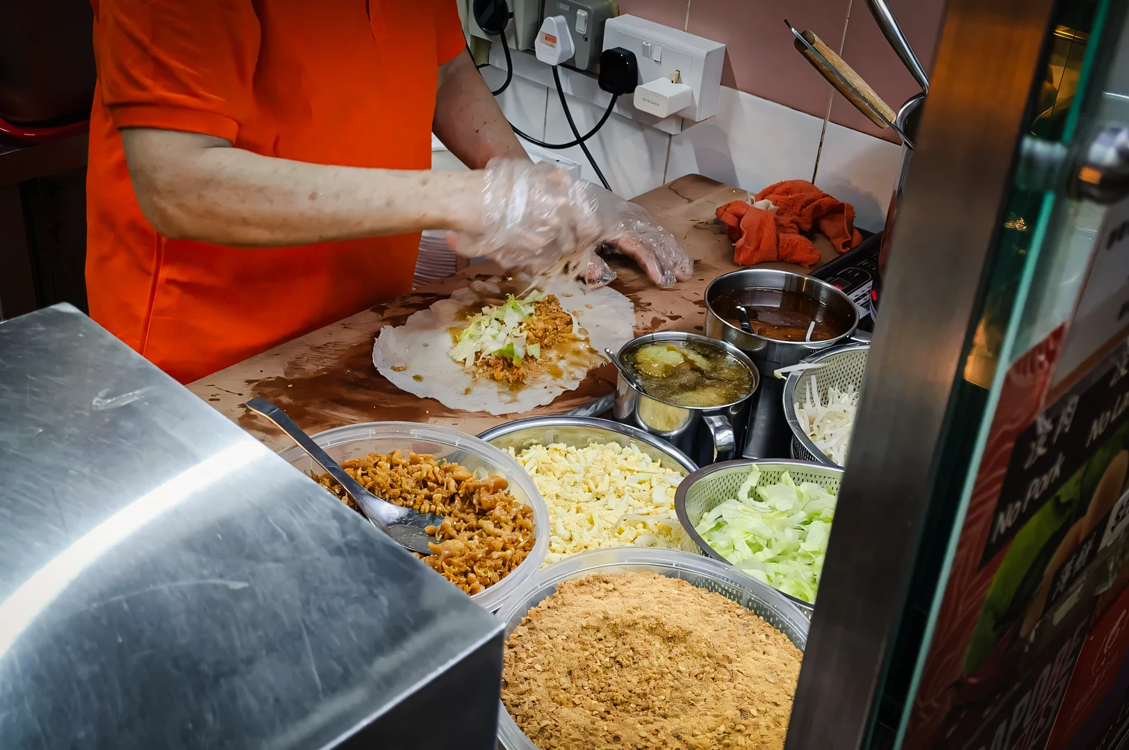 Mid-shot slightly elevated view of a food vendor assembling popiah by spreading cooked fillings, vegetables, and sauces onto a thin crepe skin at a street food stall, showing the preparation step before rolling.