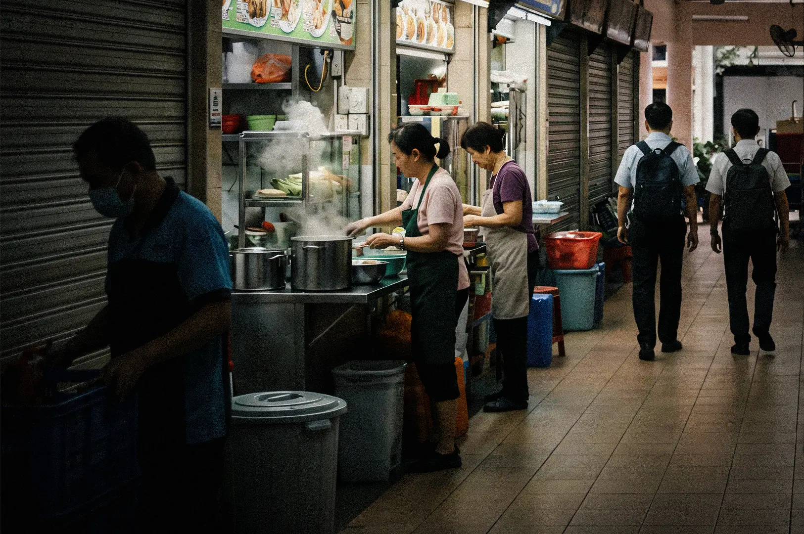 Eye‑level side‑angle wide shot of a Singapore hawker corridor where vendors prepare food at metal stalls, with steam rising from pots as pedestrians walk through the tiled walkway during morning hours.