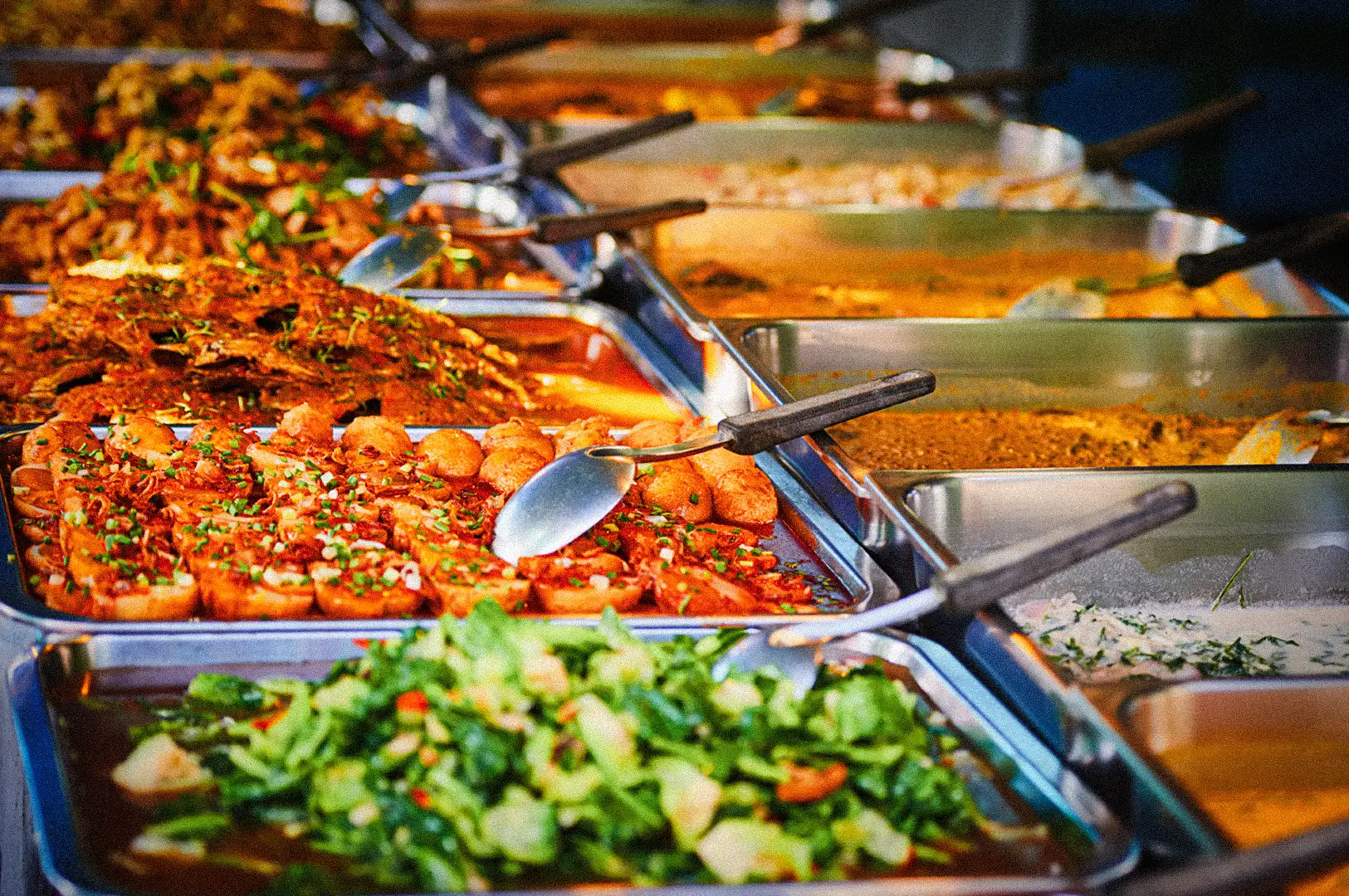 Angled close‑up buffet‑line shot of stainless steel trays filled with colorful cooked dishes, including vegetables, tofu, and sauced mains, each served with metal ladles in a cafeteria‑style food display.