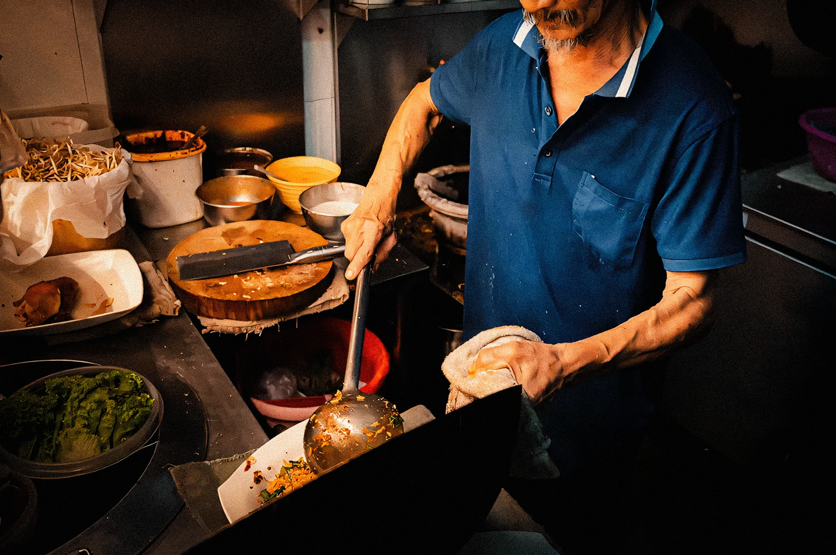 High-angle close-up of a street food vendor preparing Singaporean noodle soup, scooping broth and ingredients into bowls surrounded by stacked plates and condiments in a busy hawker kitchen.