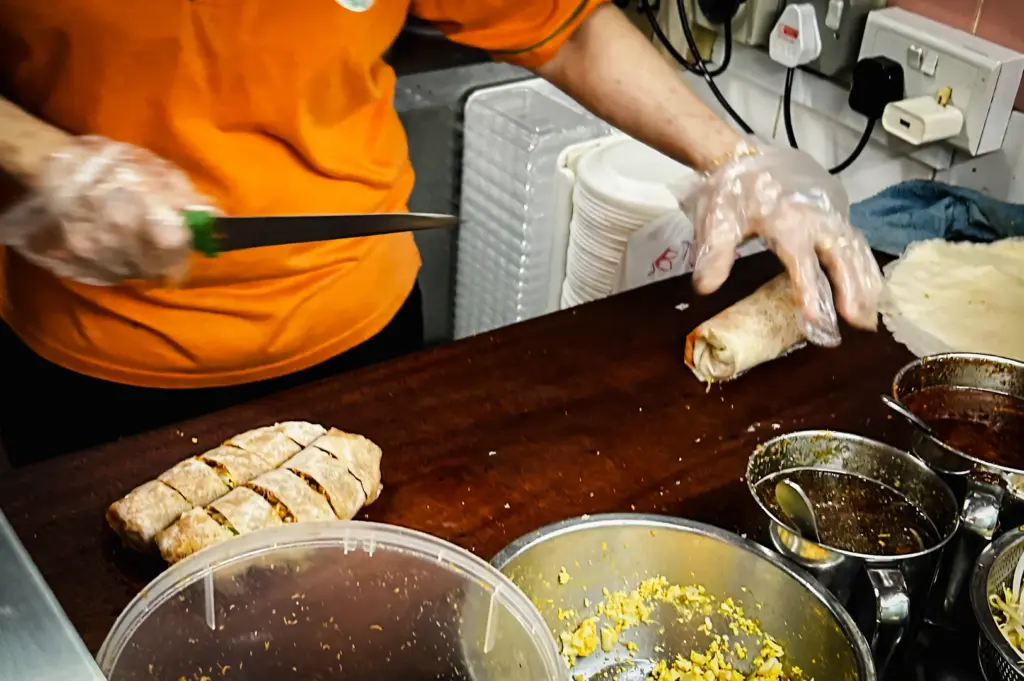 Close-up eye-level view of a vendor slicing a freshly rolled popiah into portions on a wooden counter, with bowls of fillings and sauces visible, illustrating the final step before serving.