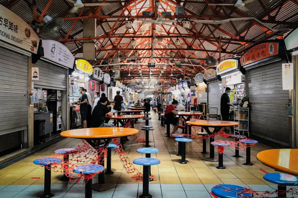 Centered eye‑level wide‑angle shot of a Singapore hawker centre in the morning, showing round tables, blue stools, partially shuttered stalls, and a few early diners having breakfast beneath a high metal roof.