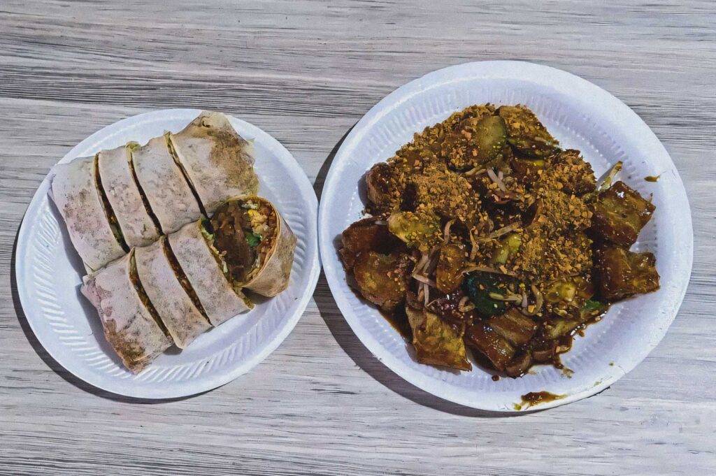 Top‑down view of sliced popiah and a plate of rojak featuring fruit, vegetables, and peanuts served on disposable plates.