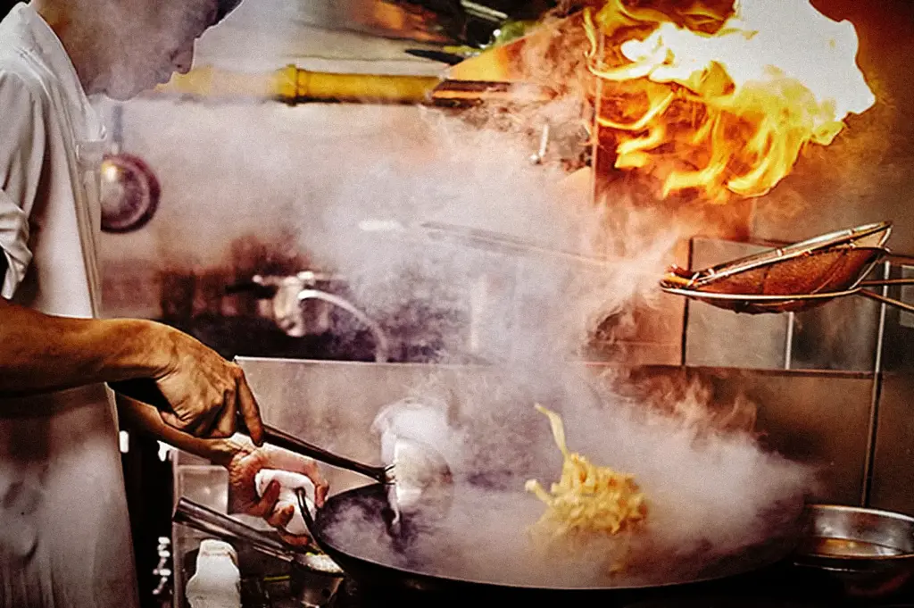 Overhead shot of a cook using wok with burst of fire and steam while sautéing noodles in high‑heat kitchen