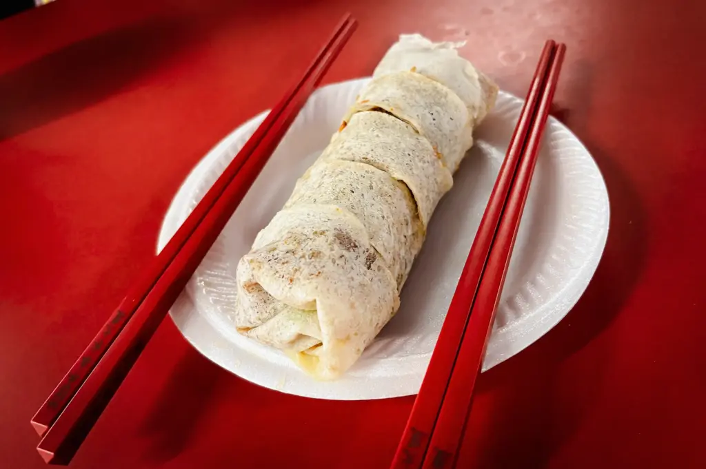 Three-quarter close-up of a sliced fresh popiah roll arranged on a white plate with red chopsticks on a red table, showing the finished Chinese spring roll wrapped in a soft crepe skin.