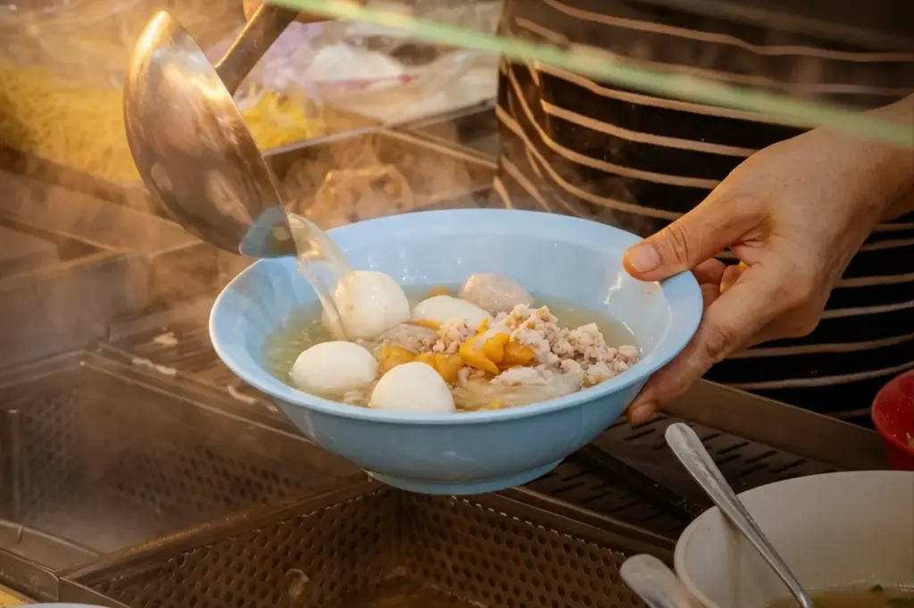 A close-up view of the final assembly of a bowl of bak chor mee at Teochew Fishball Bak Chor Mee, A hawker chef is captured pouring hot, savory broth from a large ladle into a light blue bowl filled with fresh fishballs, minced pork, and noodles, highlighting the traditional method of preparing this popular Singaporean noodle dish.