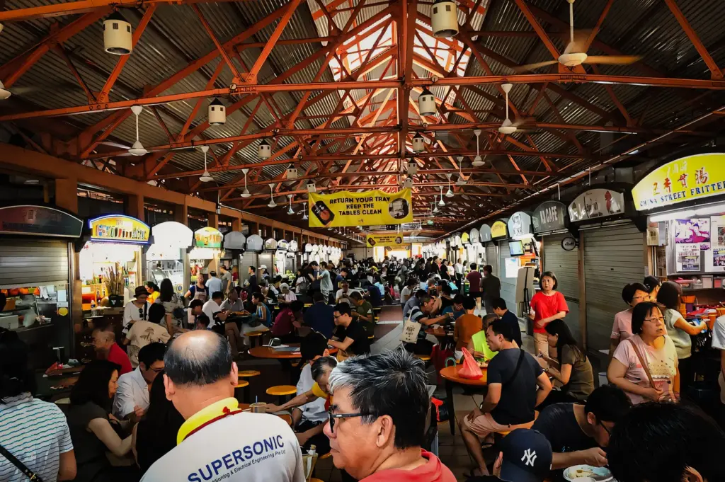 Elevated eye‑level wide‑angle shot of a Singapore hawker centre during afternoon lunch hours, crowded with diners seated at communal tables and surrounded by fully open food stalls under an orange steel roof.