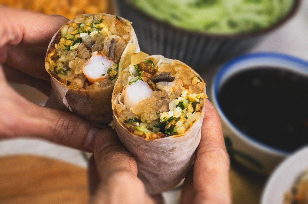 Close‑up of freshly made popiah cut in half, showing a vibrant filling of vegetables, shrimp, egg, and crispy bits wrapped in a soft spring roll skin.