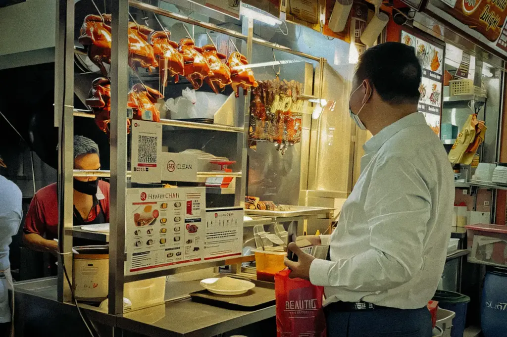 Eye‑level medium shot of a customer ordering food at a traditional hawker stall with roasted meats hanging in a glass display, menu signage visible, and warm indoor market lighting.