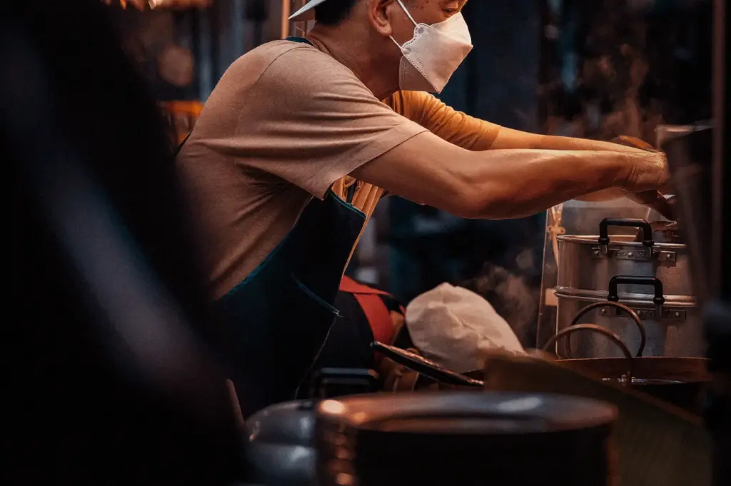 A candid moment at Teochew Fishball Bak Chor Mee capturing the routine exchange between the hawker and a customer after the food is prepared, focusing on the chef, wearing a face mask and apron, completing the service process by handing over a fresh meal and receiving payment in the fast-paced, authentic environment of a Singaporean hawker stall.