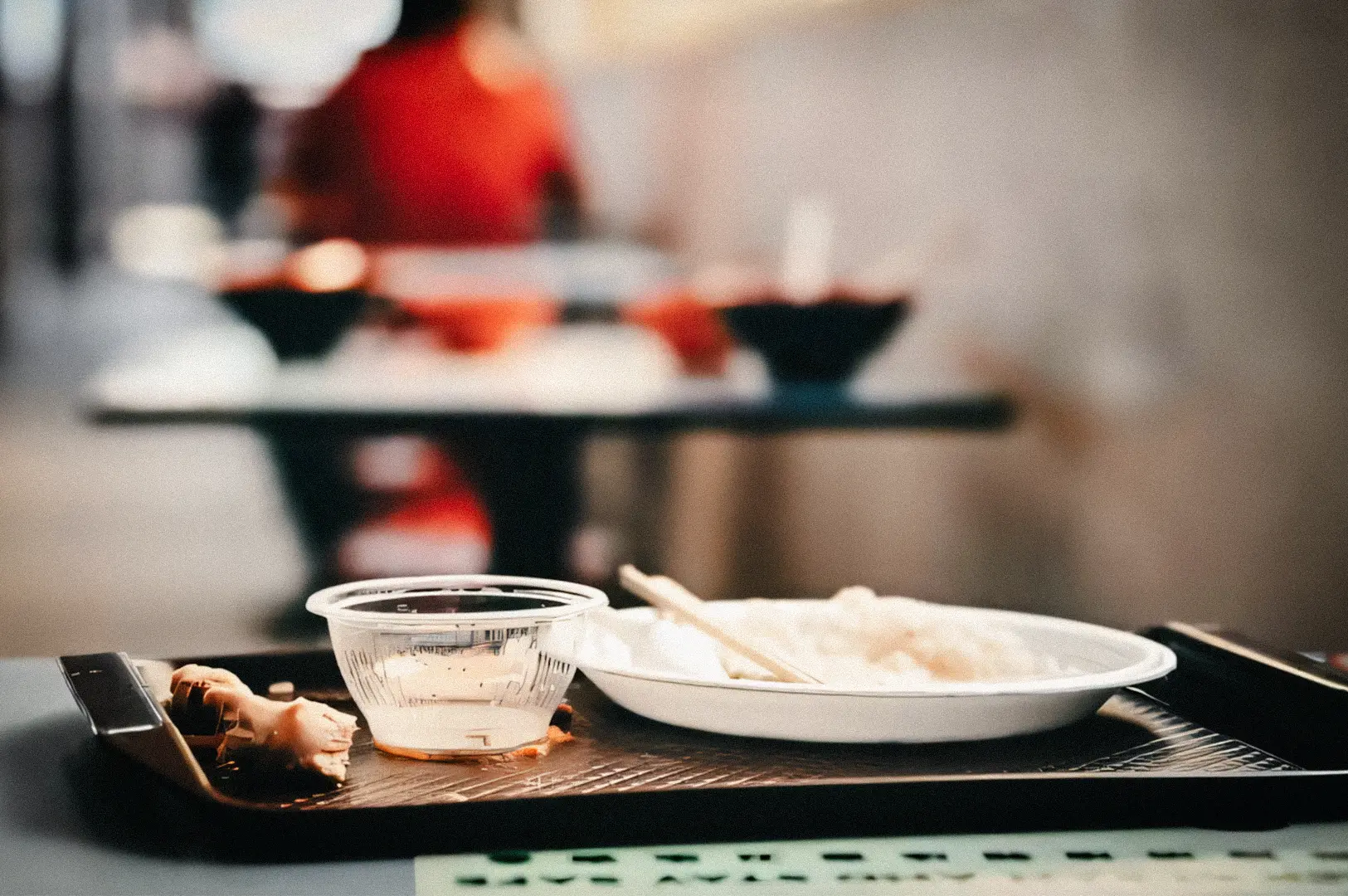 Low-angle close-up shot of an abandoned cafeteria food tray with leftover rice, chopsticks, and sauce cup on a table, softly blurred diners and seating in the background.