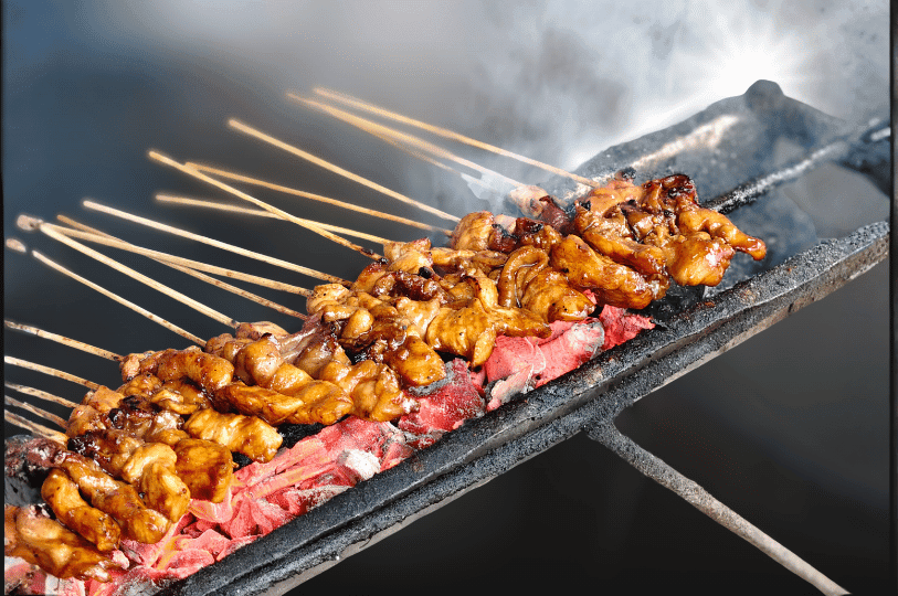Close-up of numerous marinated meat skewers cooking over glowing red charcoal, with thick smoke rising from the well-used grill.
