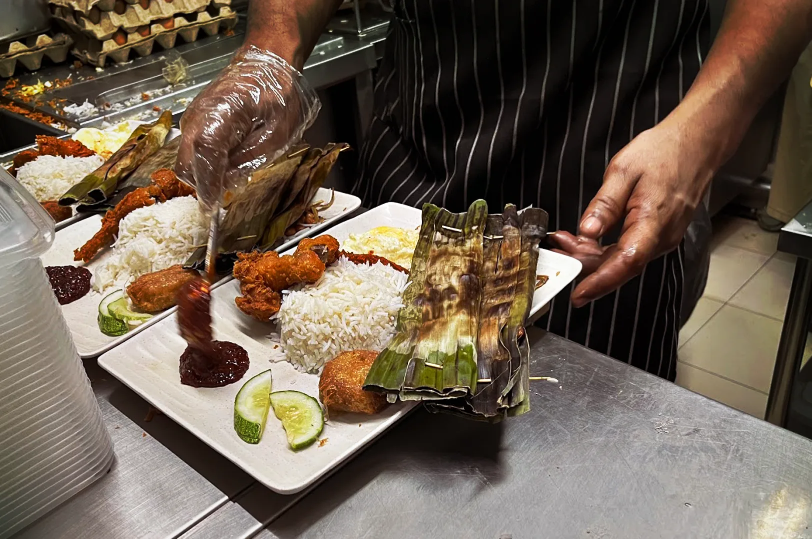 A chef plating a traditional Selera Rasa Nasi Lemak on a white plate, featuring rice, fried chicken, sambal chili, cucumber slices, and grilled otak wrapped in banana leaves.