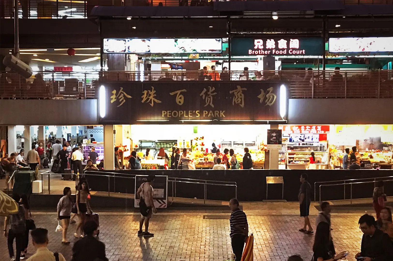Bustling night view of People's Park inside of Chinatown Complex Food Centre in Singapore. The vibrant hawker center exterior features glowing neon signs in English and Chinese for restaurants, and busy hawker food stalls, with tourists and locals walking in the foreground marketplace.