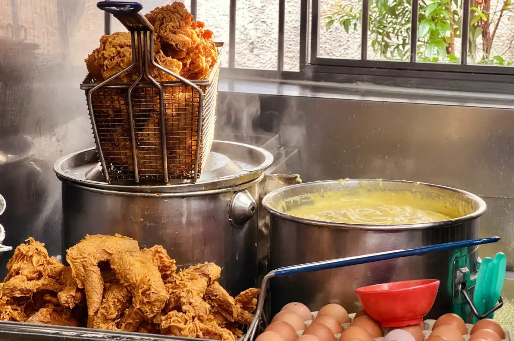 Freshly fried crispy chicken wings, raw eggs, and a large pot of steaming gravy at a Selera Rasa Nasi Lemak stall, highlighting the high-quality ingredients used in their famous Singaporean dish.