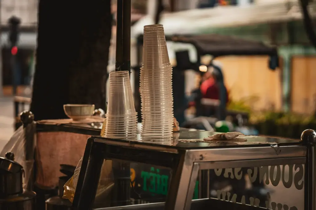 A quiet, still scene at a hawker centre food stall before the rush, featuring clean, stacked plastic cups on a metal counter awaiting the first patrons of the day.