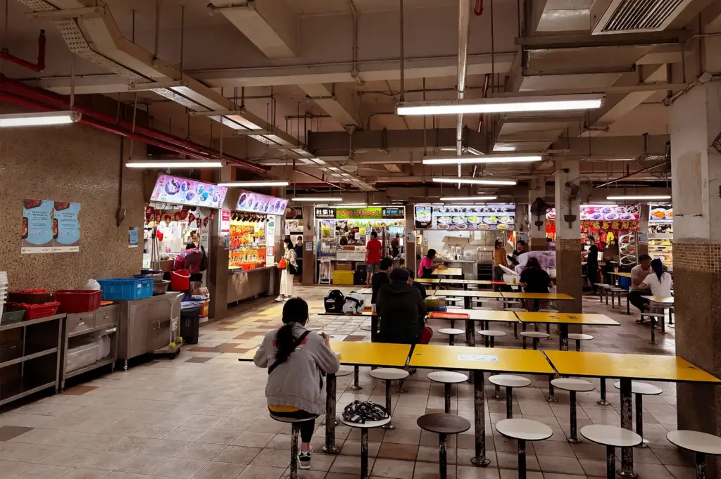 Grid-like architectural structure of Singapore's Chinatown Complex Food Centre hawker center, featuring symmetrical rows of yellow tables, overhead fluorescent lighting, and local food stalls running continuously without break.
