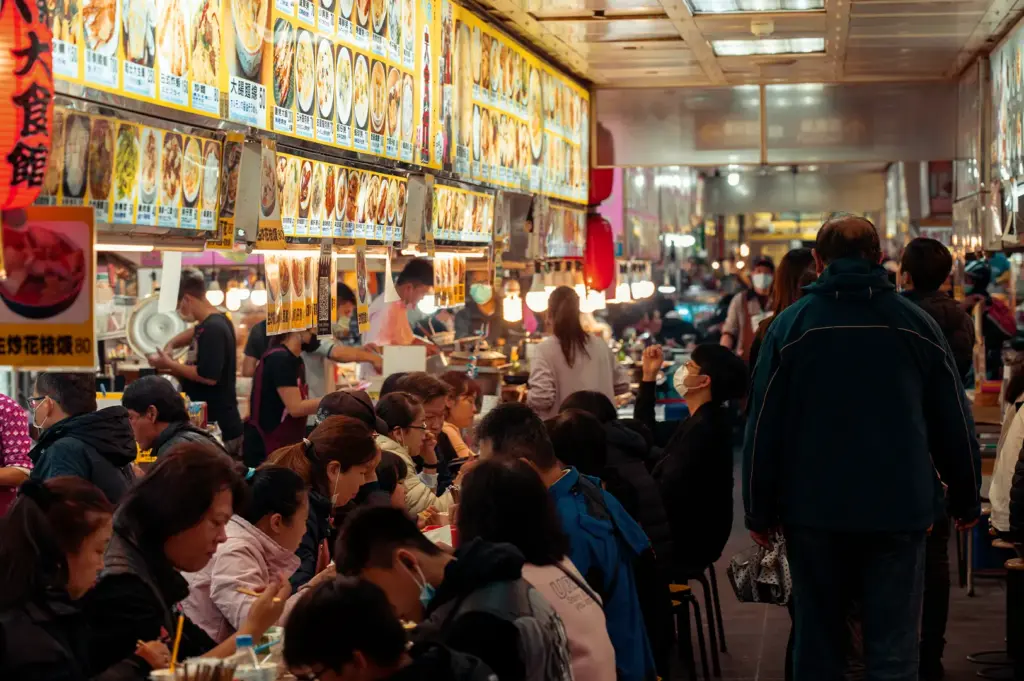 The transition from preparation to peak dining hours at a crowded hawker centre, showcasing the vibrant atmosphere where rows of patrons enjoy meals beneath extensive overhead menus.