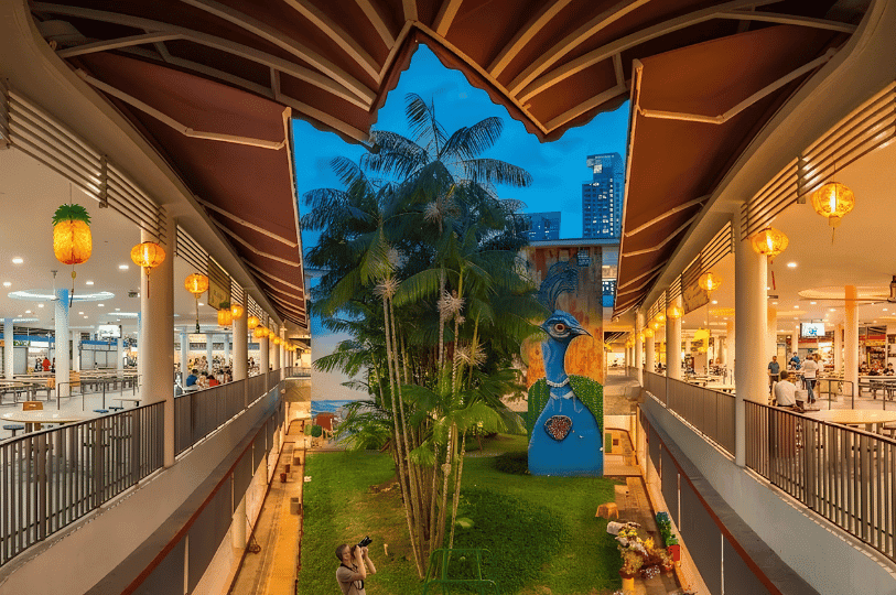 A broad view of Tiong Bahru Market’s central courtyard under cool daylight, highlighting the layered architectural design that separates the active upper dining corridors from the ground-level green space. The open-air structure frames a tall peacock mural, demonstrating how the market balances structured pedestrian flow with an airy, communal environment.