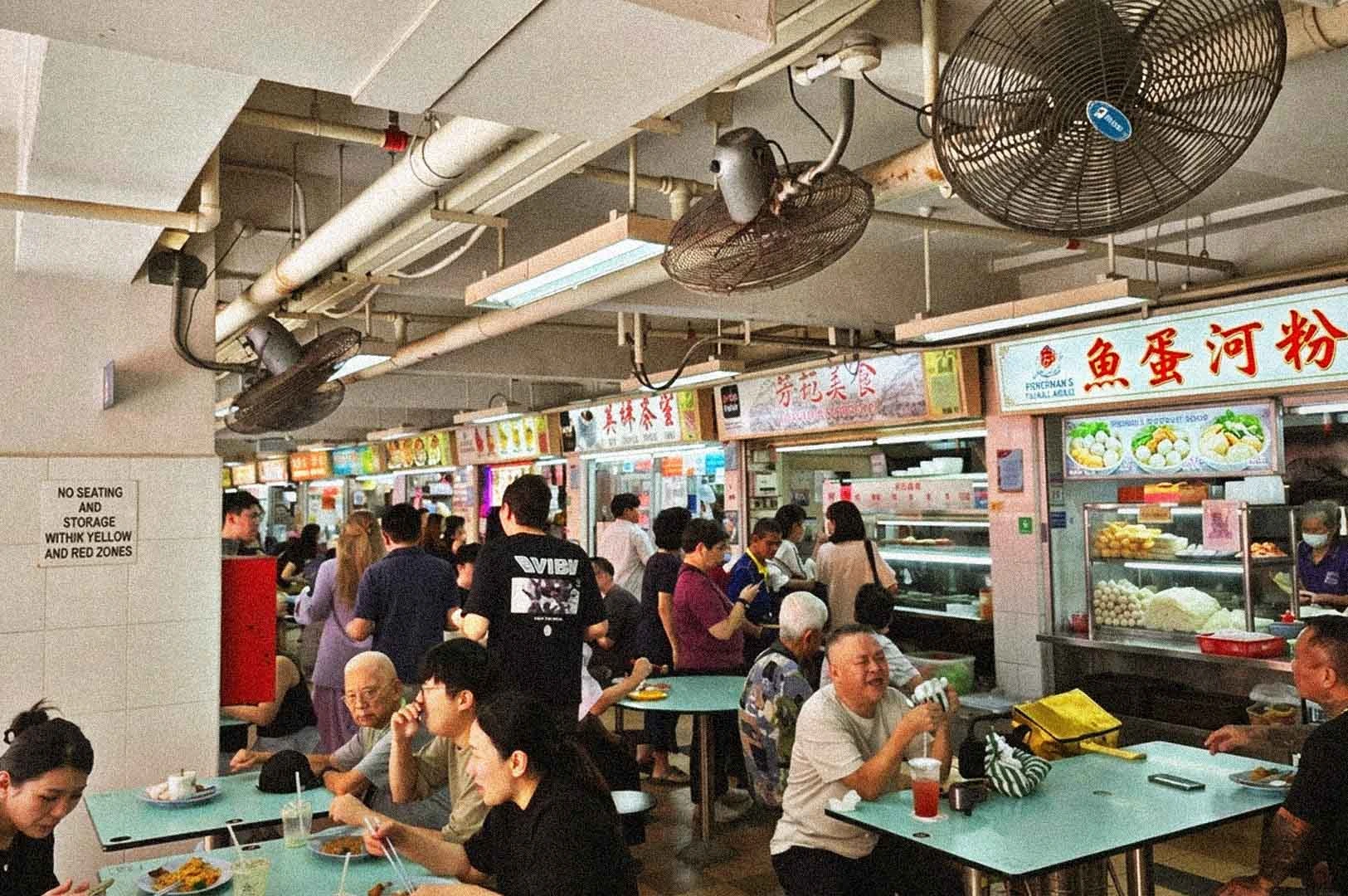 Busy lunchtime crowd at Hong Lim Food Centre in Singapore featuring hawker stalls like Fishball Noodles stalls, patrons dining at turquoise tables, and overhead fans.