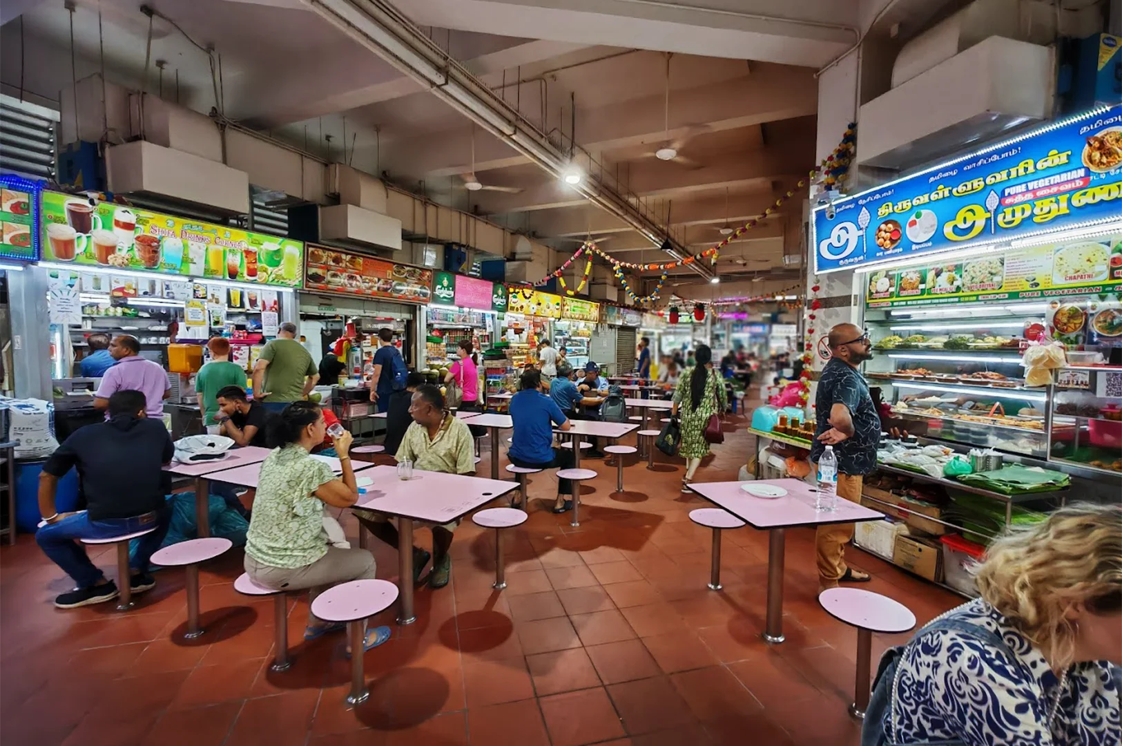 Inside a bustling Singaporean hawker centre with tiled floors, patrons dine at simple tables while diverse food stalls display menus, highlighting the enduring community atmosphere of traditional open-air dining.