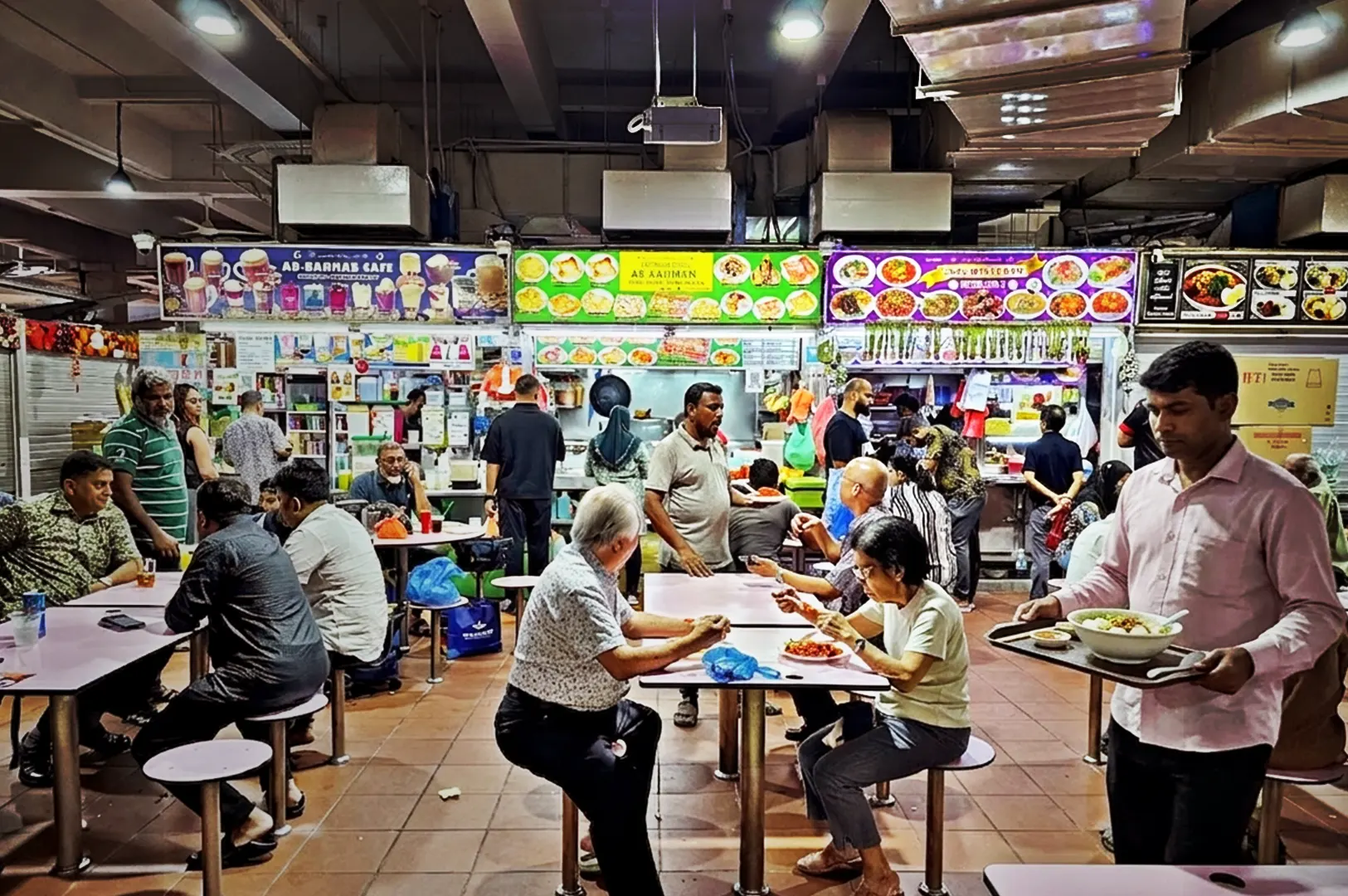 Busy indoor hawker food center in Singapore featuring patrons eating at tables and a man in a pink shirt walking while carrying a tray with a bowl of fishball noodle soup.