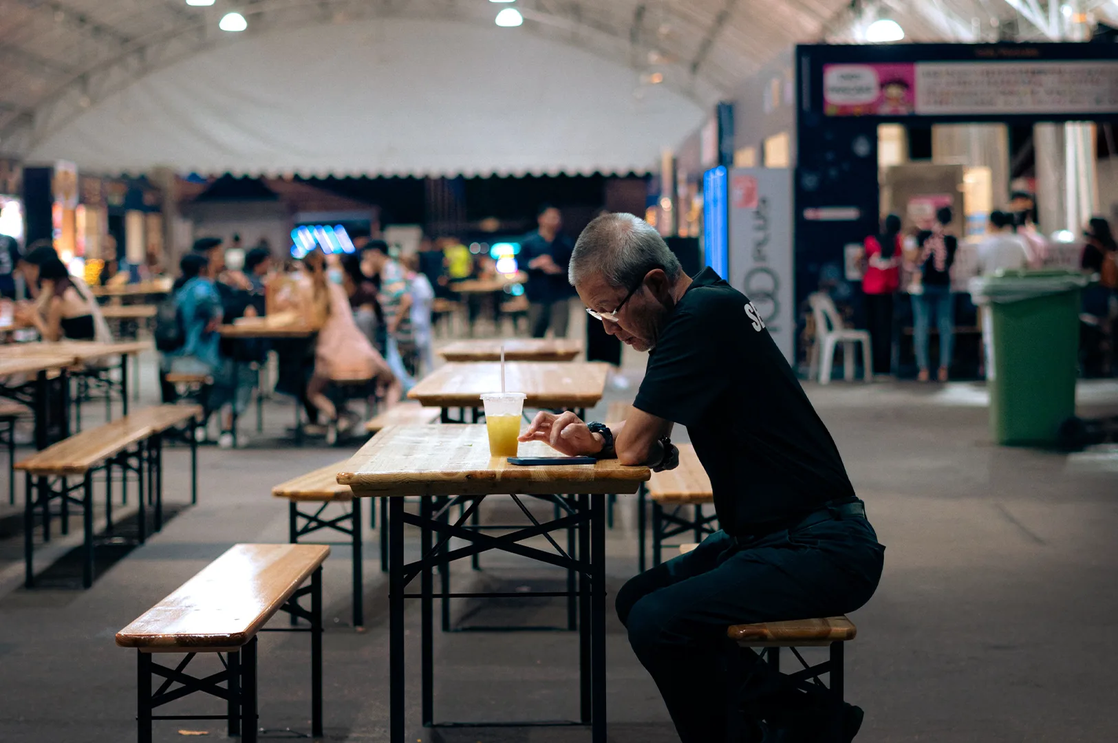 A man in a black shirt sits alone at a wooden table in a busy Singaporean hawker centre looking at his smartphone while patrons eat and walk through the background.