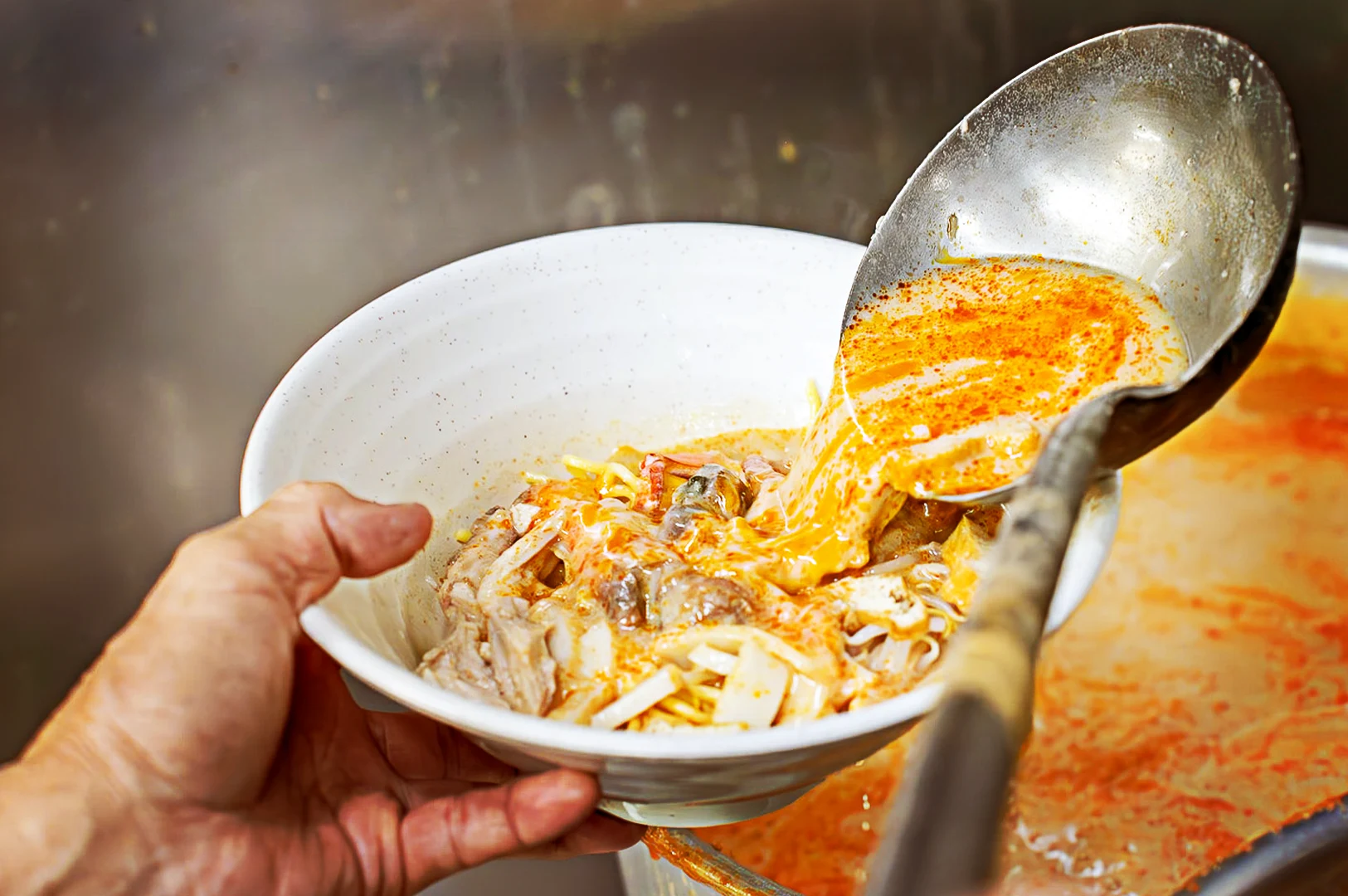 Laksa broth being ladled into a bowl filled with noodles, meats, cockles, for the final assembly of an authentic Laksa dish.