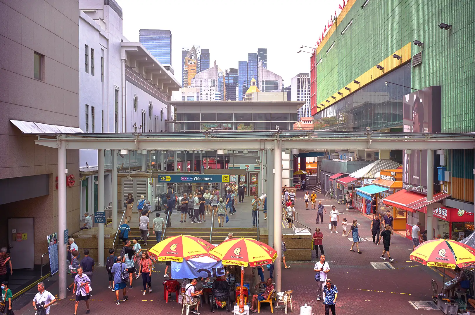 Pedestrians walking through an outdoor plaza near the Chinatown MRT station entrance in Singapore during the busy lunch rush hour, surrounded by urban buildings and food stalls with colorful umbrellas.