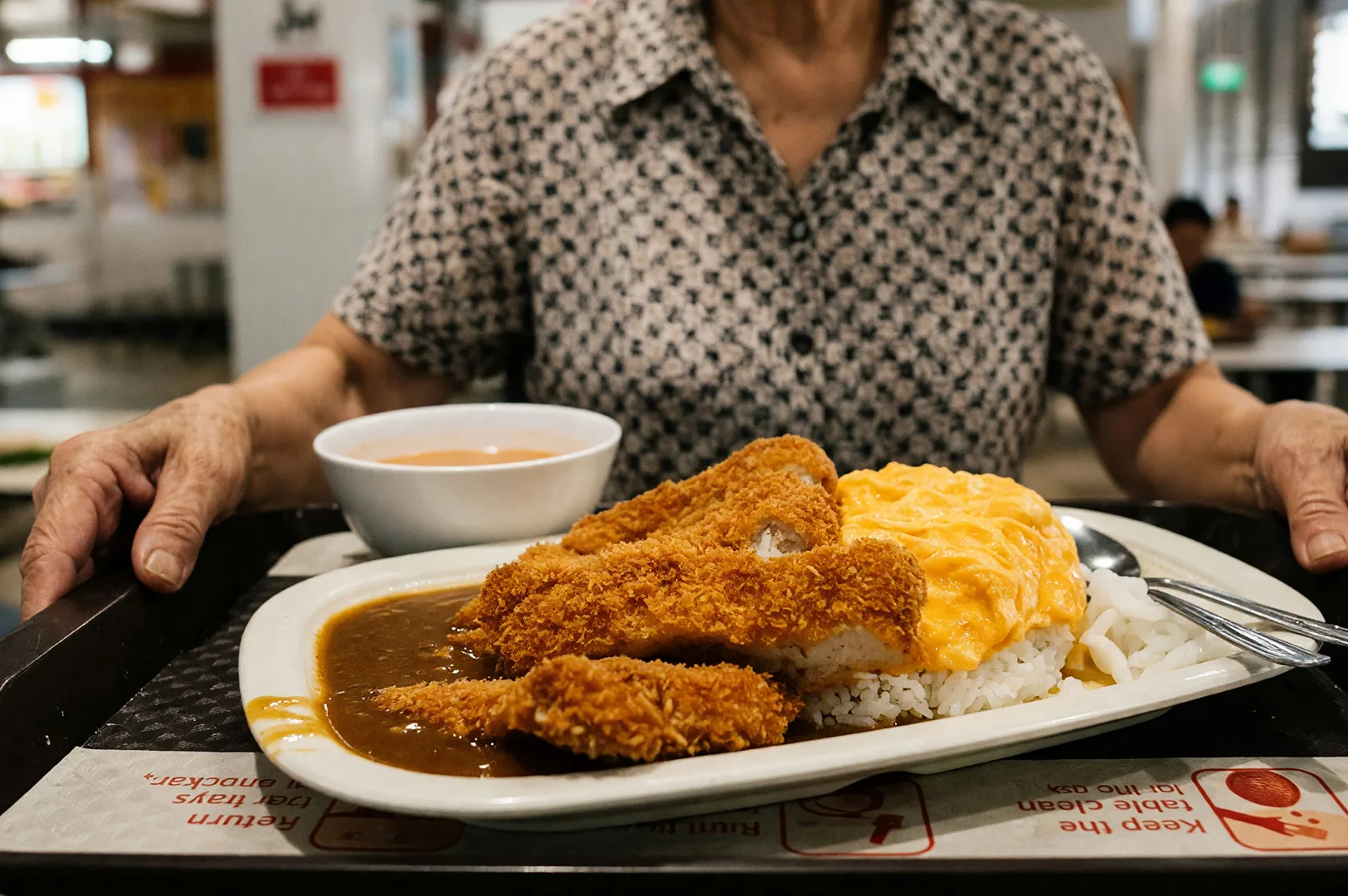 A diner carrying a plate of Japanese curry on a tray down the aisle of Tanjong Pagar Plaza Market & Food Centre in Singapore, surrounded by seated patrons