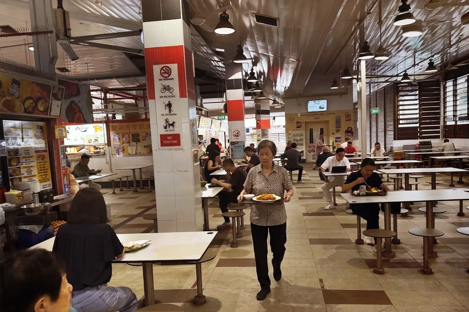 A diner carrying a plate of Japanese curry on a tray down the aisle of Tanjong Pagar Plaza Market & Food Centre in Singapore, surrounded by seated patrons