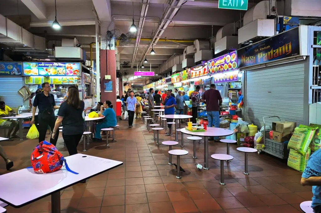 A view of a traditional, older Singaporean hawker centre featuring rows of food stalls with rolled-down shutters, diners at circular tables, and a utilitarian ceiling structure, reflecting the authentic and lived-in environment of local food culture.