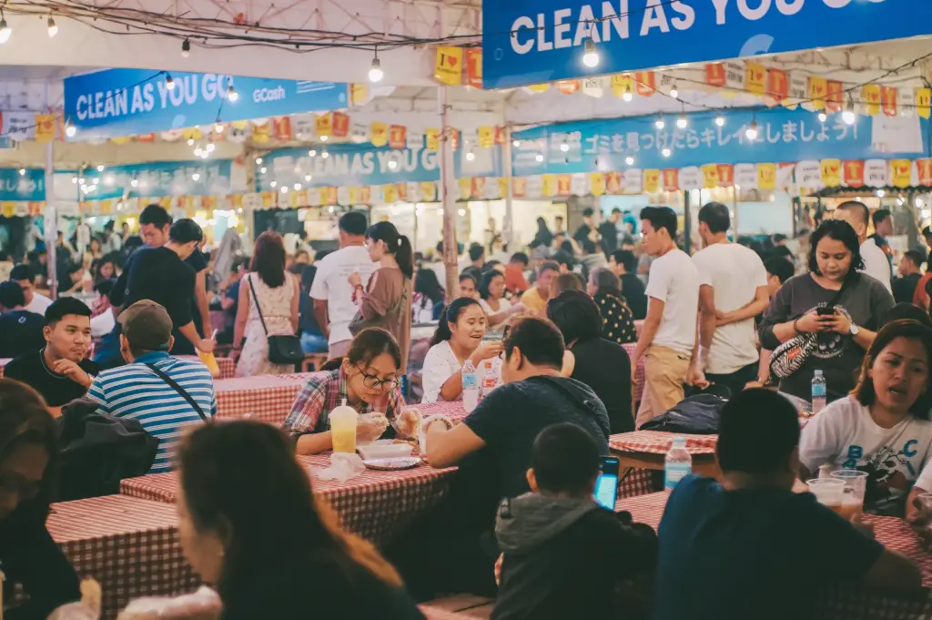 A crowded hawker center during the peak lunch rush, featuring people sitting at red-and-white checkered tables eating meals under "Clean As You Go" banners and decorative bunting.