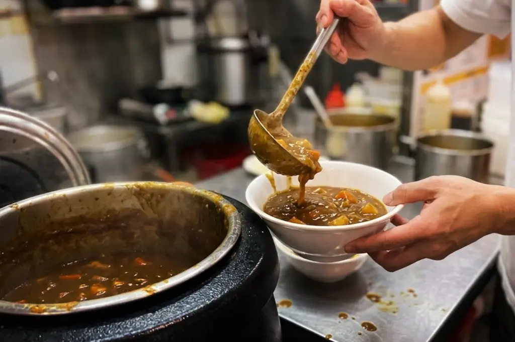 A chef ladling rich, thick Japanese curry with potatoes and carrots into a white bowl at a hawker stall in Tanjong Pagar Plaza Market and Food Centre.