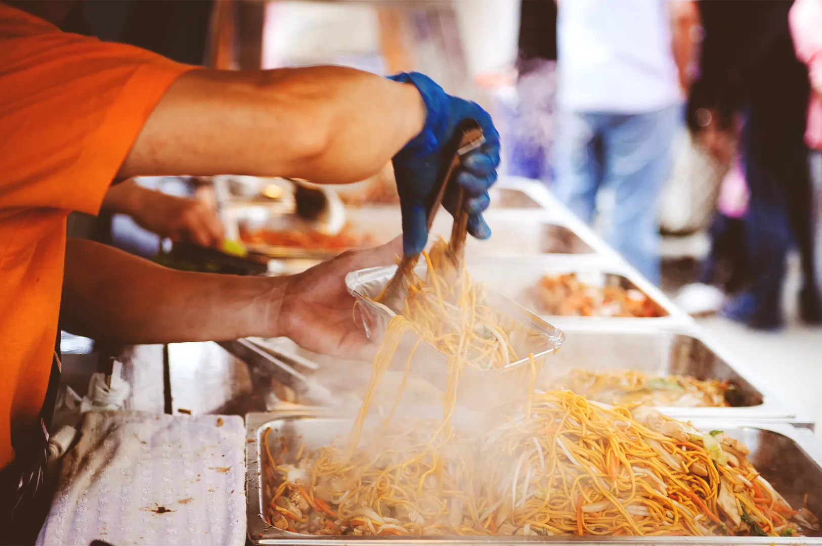 A street food vendor in an orange shirt uses tongs to pack steaming stir-fried noodles into a takeaway container, demonstrating the repetitive, efficient sequence used to maintain food quality during a busy lunch rush.