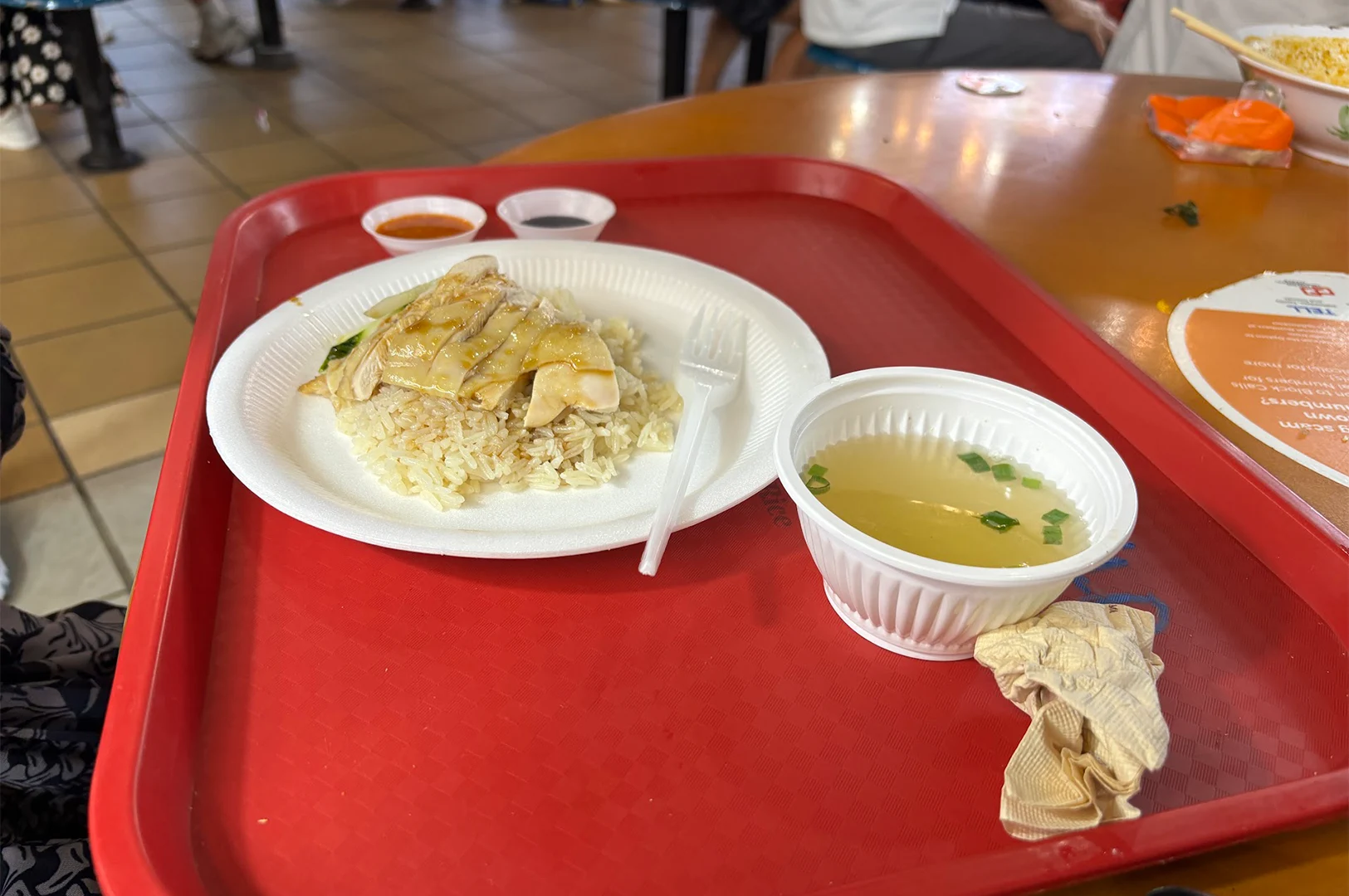 A serving of Hainanese chicken rice on a red plastic tray at Maxwell hawker centre, showcasing the worn surface of the tray that reflects years of repeated daily use in a bustling food market.
