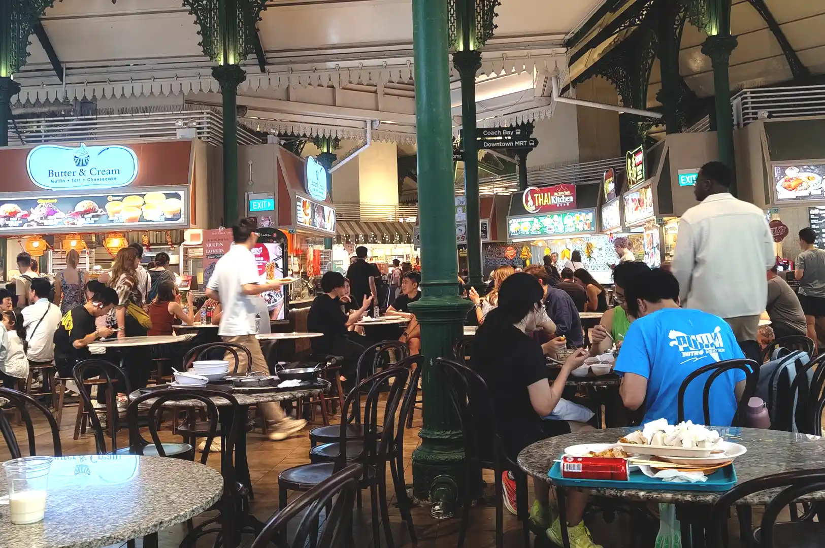 A bustling scene at Lau Pa Sat food center in Singapore, showing patrons dining at marble-topped tables, with various food stalls visible in the background.