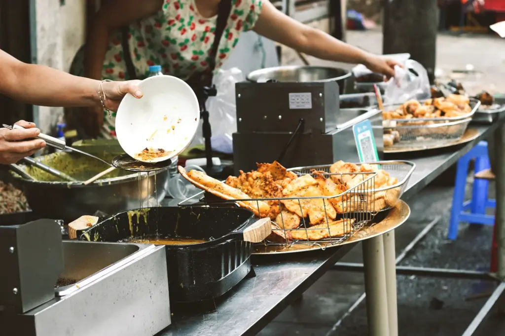 A food stall worker pours batter into a hot pan alongside baskets of pre-fried snacks, showcasing the disciplined and consistent preparation method that remains unchanged despite the fluctuating demand of a lunch