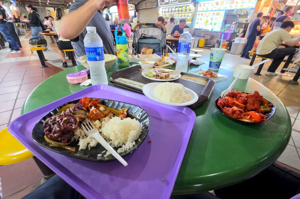 A busy circular table at Maxwell food centre crowded with multiple plastic trays and half-eaten meals, highlighting how the surfaces and shared dining tools carry quiet marks of constant, daily wear.