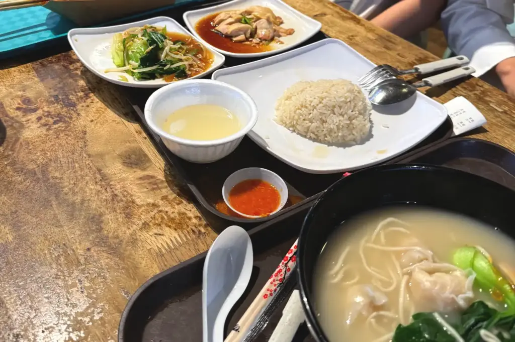 A close-up of a spread of traditional Singaporean hawker dishes on a wooden table, including chicken rice, vegetable sides, and wonton noodle soup, capturing the essence of local dining culture.