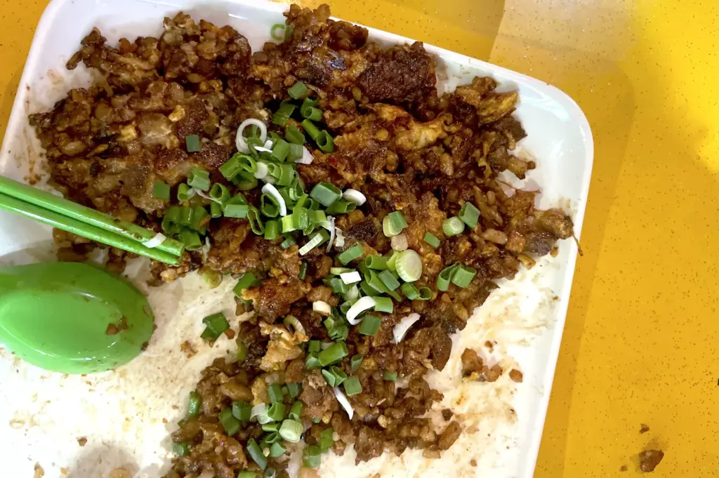 Half-eaten plate of black carrot cake with green chopsticks and a spoon, set against a classic yellow hawker table, depicting the authentic dining experience and everyday routine at Lau Pa Sat.