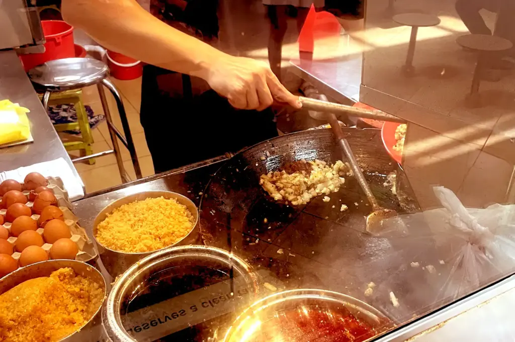 A hawker in motion behind a glass partition, expertly using a spatula to stir-fry char kway teow in a hot wok, capturing the daily culinary routine behind a popular Singaporean stall.