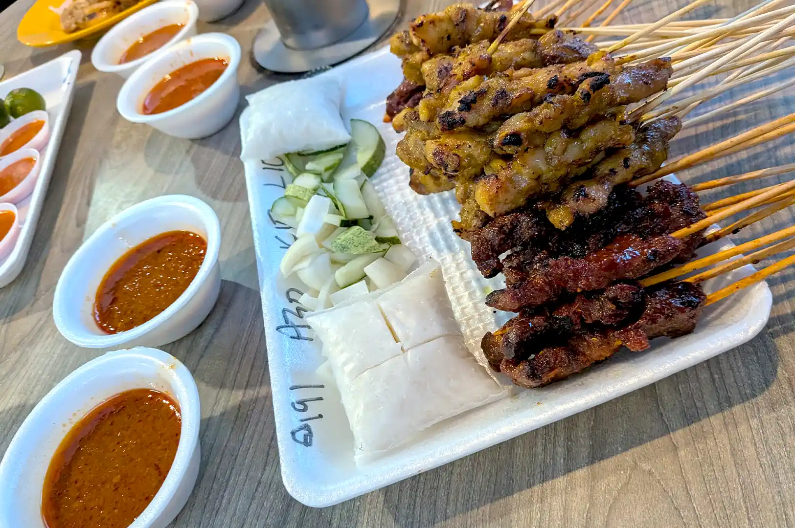 A white styrofoam tray piled high with grilled meat satay skewers alongside cucumber slices, onions, rice cakes, and small bowls of peanut dipping sauce at East Coast Lagoon Food Village.