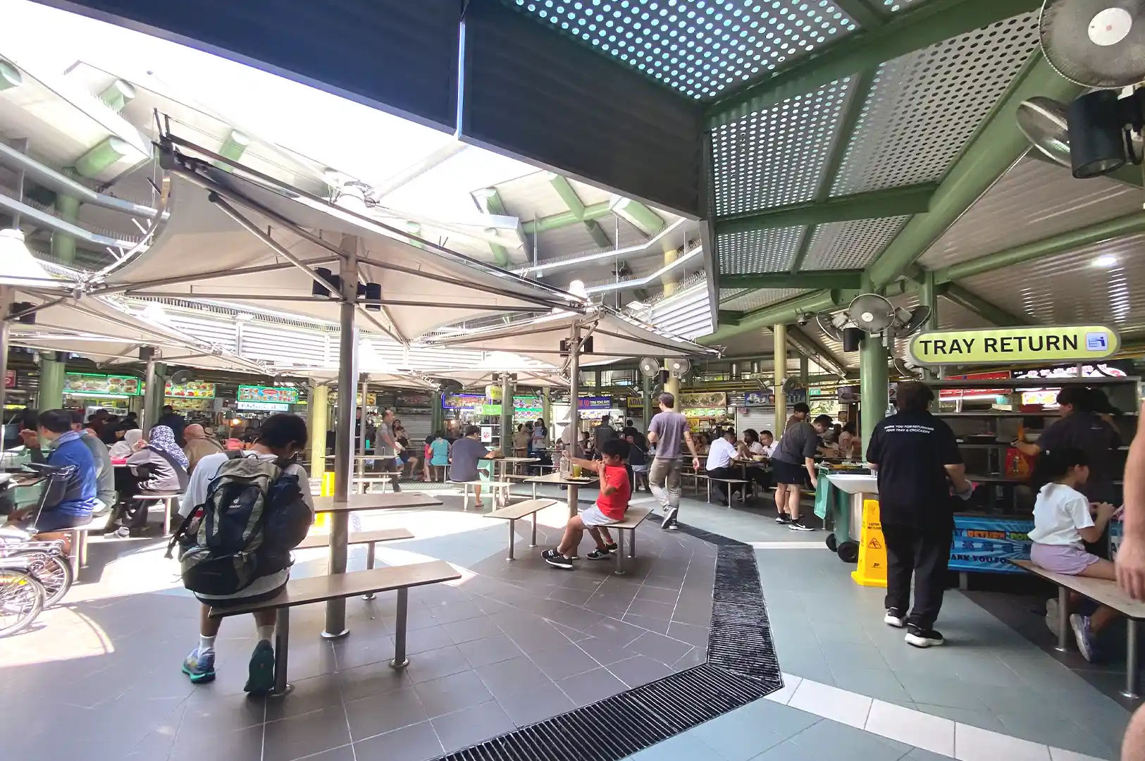 A wide-angle view of the bustling Adam Road Food Centre featuring high-roof awnings, shared tables, and patrons moving through the open-air hawker center.
