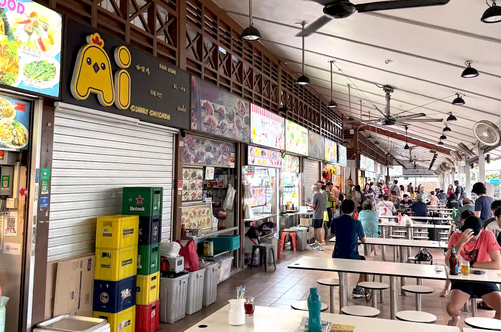 A bustling open-air hawker center with rows of food stalls, stacked crates, and diners seated at tables beneath ceiling fans during busy operational hours.