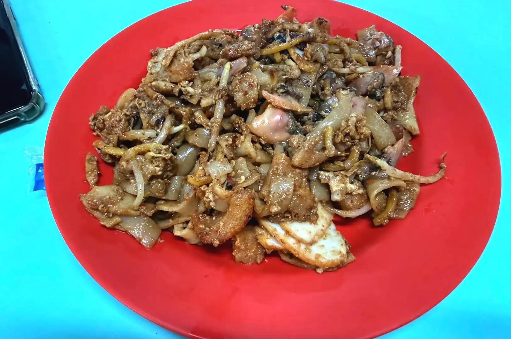 Close-up of glossy char kway teow on a bright red plate at Hong Lim Food Centre, capturing the glistening surface of flat noodles and cockles fried in wok hei.
