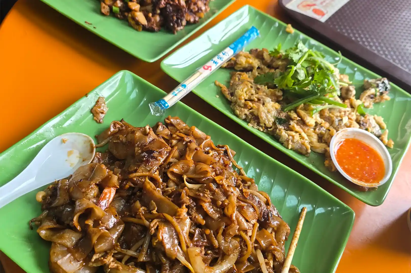 Green plates of fried local hawker noodles and oyster omelet served on a bright table, reflecting the cycle of preparation and cleaning that defines a hawker stall's day.